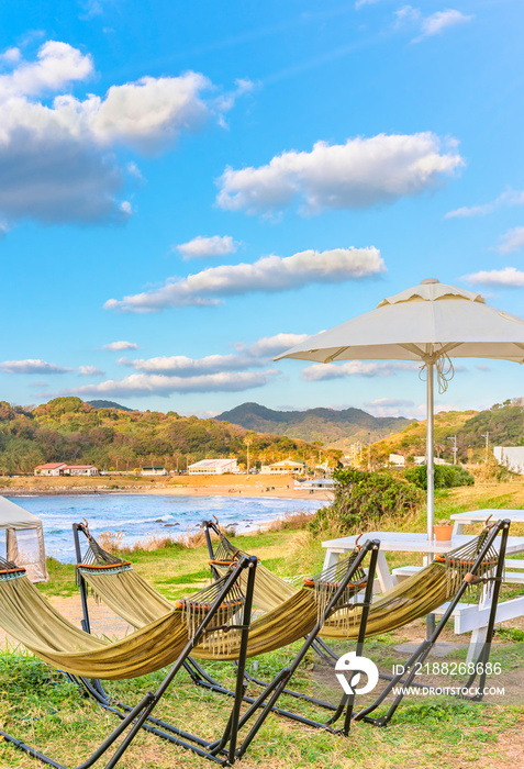 Hammocks and wooden outdoor tables with benches overlooked by a parasol  along the coast of Itoshima beach at sunset close to Sakurai Futamigaura’s Couple Stones of Fukuoka in Japanese Kyushu island.