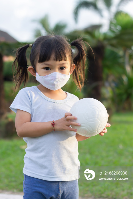 Vertical Portrait image of 2-3 years old baby. Happy​ Playful Asian child girl weraing fabric face mask. She play the ball toy at the green park playground. Kids and healthy. Covid-19 outbreak.