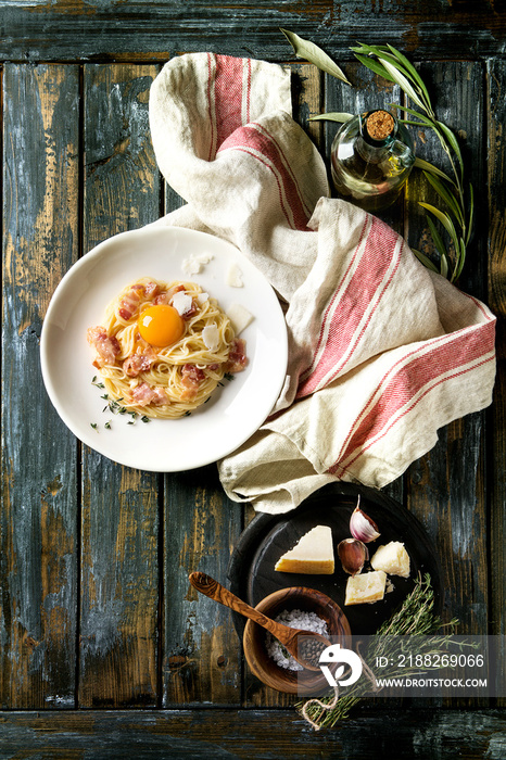 Italian traditional pasta alla carbonara with yolk, pancetta bacon, parmesan cheese, thyme, served in white plate on textile over old wooden plank background. Ingredients above. Top view with space