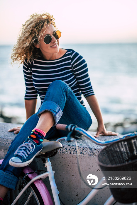 Happy beautiful blonde long curly hair woman enjoy the sunset sit down with blue ocean and sky in background - outdoor leisure activity for young pretty active people