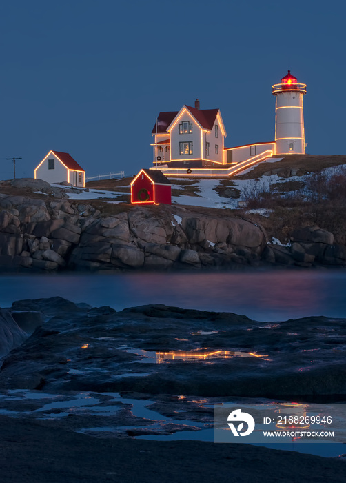 Nubble Lighthouse (also known as Cape Neddick Lighthouse) located in York, Maine with Christmas lights and a reflection in a pool of water.