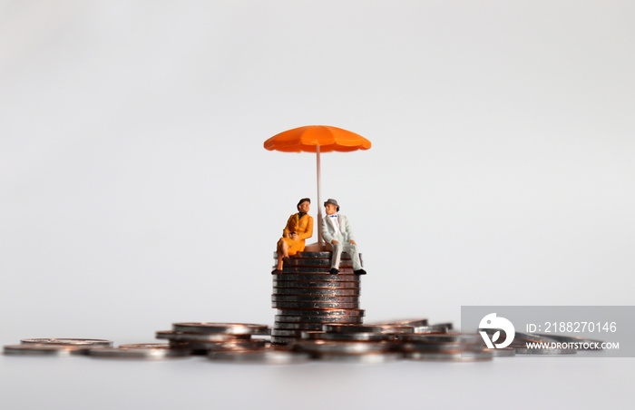 An elderly miniature couple sitting with an umbrella on a pile of coins.