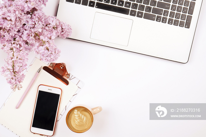 Feminine desktop, close up of laptop keyboard, blank paper sheet on clipboard, coffee, smartphone, lilac flower. Notebook computer, blank screen cellphone, cappuccino cup, white background. Copy space