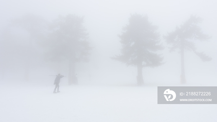 Silhouette of a man walking with skiis on a shoulder in extreme fog against barely visible trees