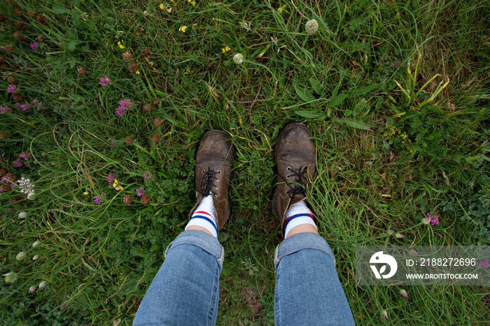 Top shot of hard brutal leather boots and legs wearing denim jeans, stand in middle of grass field full of flowers