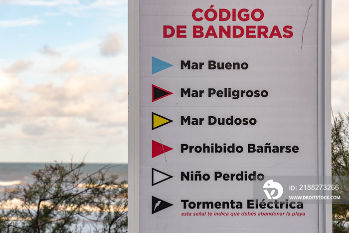 Indicative sign of the codes of the flags that announce the dangerousness of the sea. Written in Spanish in its entirety. With a background of bushes, beach and a sky with some clouds