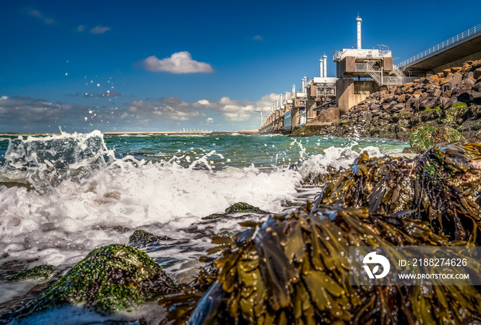 View on the architecture of the flood barrier ’Neeltje jans’ in the Netherlands from the rocky coast with seaweed and splashing waves