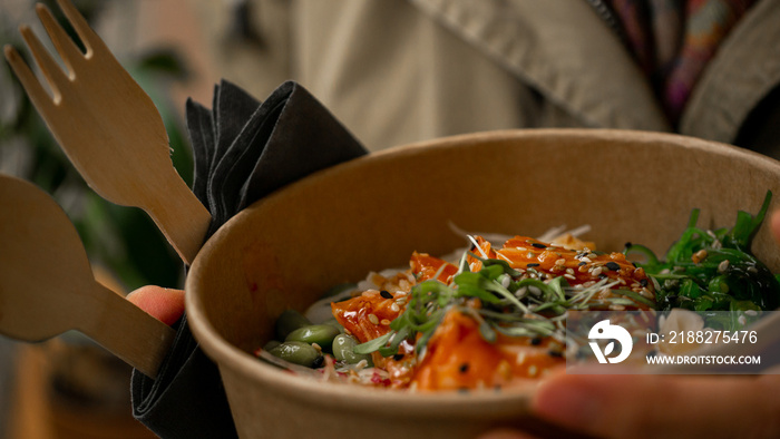 Female enjoying quick gourmet lunch - plastic-free, recyclable take away bowl with raw fish, rice and vegetables Japanese style. Macro, differential focus, close up.