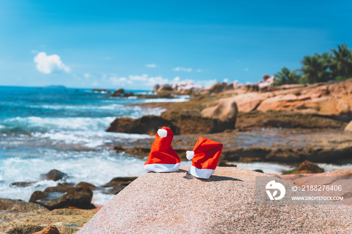 Couple of santa claus hat on tropical exotic paradise sandy beach with ocean waves and rocky coastline in background