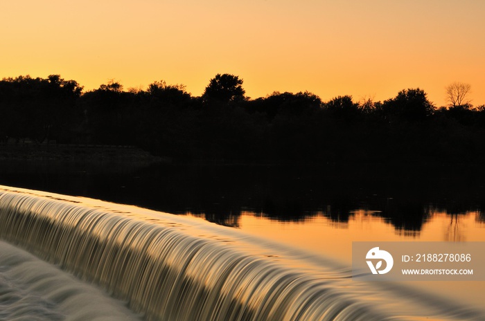 Tranquil water turns to white water as it flows over a dam in the Fox River in South Elgin, Illinois while reflecting the the glow from the setting sun beyond the trees on the far bank.