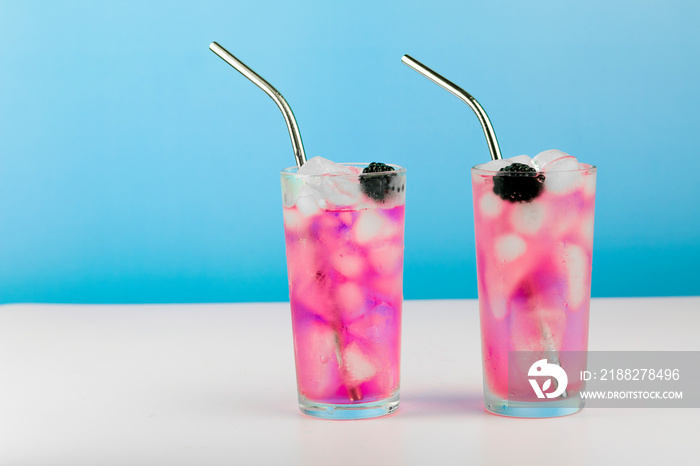 Summer refreshing pink cocktail with ice cube, metal straw and blackberry on blue background.