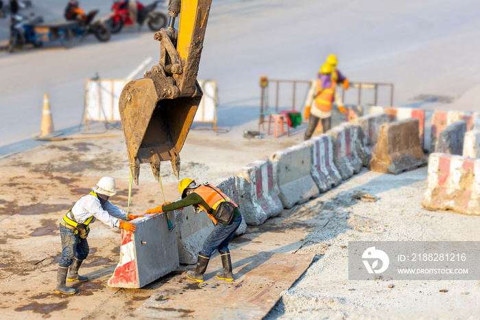 Tow man workers wear safety suits helped to lift and arrange the cement concrete barriers by stick boom arm of excavator In harmony and actively