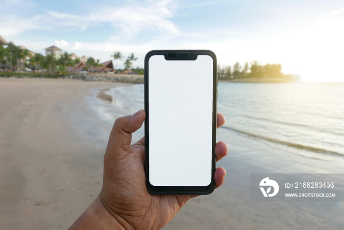 Close up of hand holding phone with white screen by the beach. Smartphone with mockup on background beach and hotel or resort.