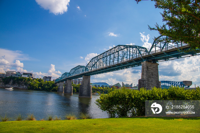 the Walnut Street Bridge over the rippling blue waters of the Tennessee river with rocks along the banks surrounded by lush green trees and grass with blue sky and clouds at Coolidge park