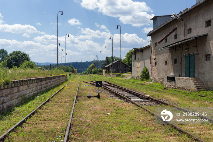 Abandoned and neglected train station in small town. Old rusty rails overgrown with green vegetation.