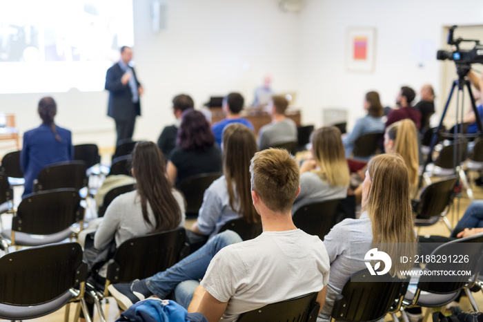 Speaker lecturing in lecture hall at university. Students listening to lecture and making notes.