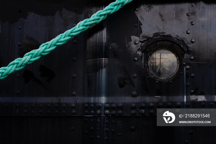 Black ship hull texture background. Rusty detail of dark, weathered marine tanker bow with rivets, green rope and porthole. Low-key, scenic, cinematic, fairy, historical, moody backdrop. Copy space