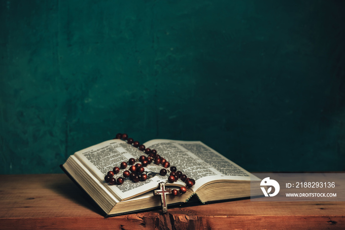 Open Holy Bible and beads crucifix on a red old wooden table. Beautiful green wall background.