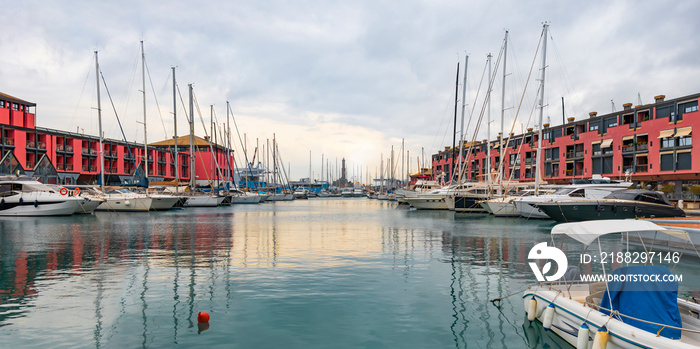 Panoramic view of moored yachts in the port of Genoa, with a lighthouse in the background, yachts and buildings reflected on the smooth surface of the water