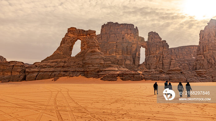 Tamesguida cathedral in Tadrart Rouge. Walking tourists. Five silhouettes with shadows walking away, orange color sand with sunny cloudy sky. Naturally sculpted rocky mountains of the Tassili N’Ajjer