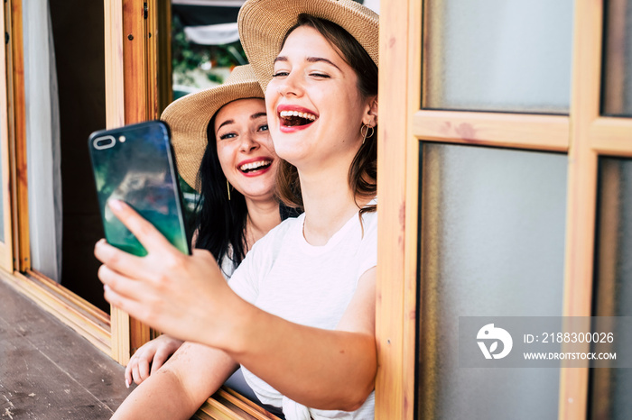 Happy couple of young woman caucasian friends have fun together at the window enjoying the phone int