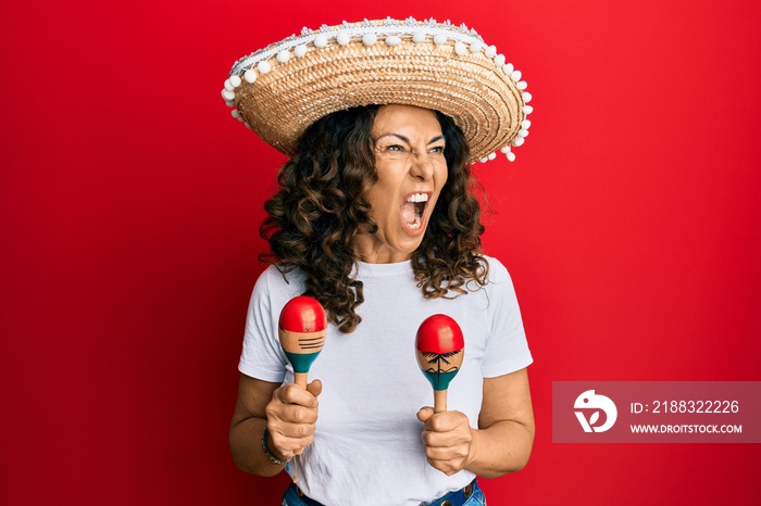 Middle age hispanic woman holding mexican hat playing maracas angry and mad screaming frustrated and