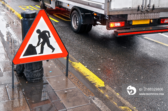 人行道旁潮湿道路上的道路工程警告标志，在公共汽车站上方背景中行驶的面包车写着