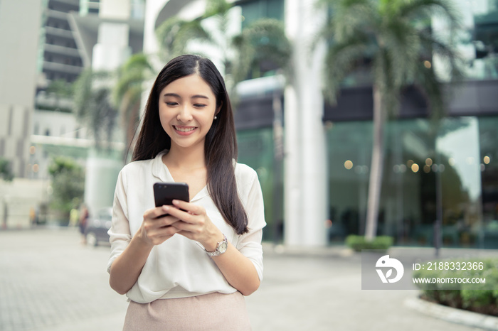 年轻的亚裔中国女商人微笑着在S办公楼附近的户外使用数字智能手机