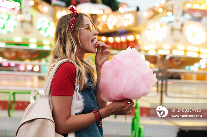 Image of pretty cute woman eating sweet cotton candy while walking in amusement park