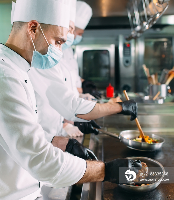 Chefs in protective masks and gloves prepare food in the kitchen of a restaurant or hotel.