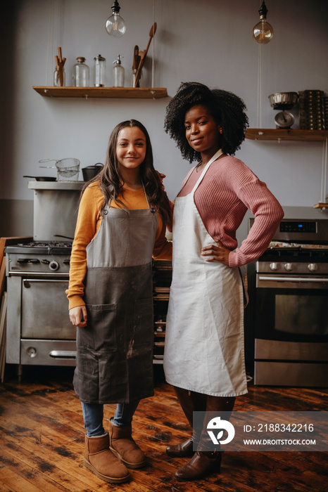 Portrait of smiling female chef with teenage student wearing aprons while standing against wall in c