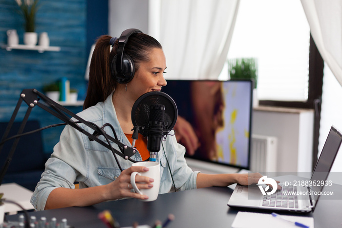 Woman vlogger holding cup and streaming podcast using laptop. Influencer with headphones in home off