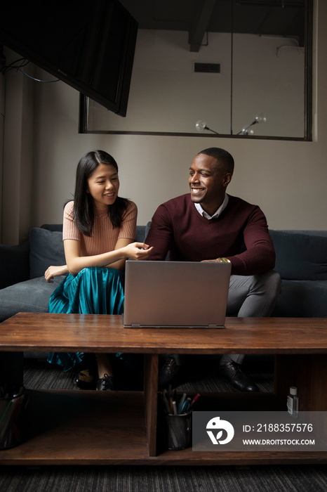 Smiling business people talking while sitting by laptop computer on sofa against wall in office