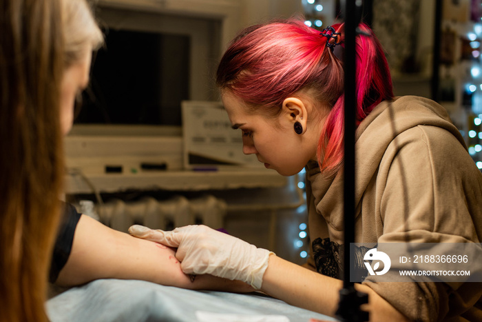 Charming girl with pink hair and tattoos stuffs a tattoo on a womans hand in a tattoo parlor, creat