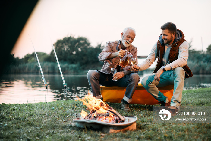 Happy senior fisherman and his son drink wine by campfire at sunset.