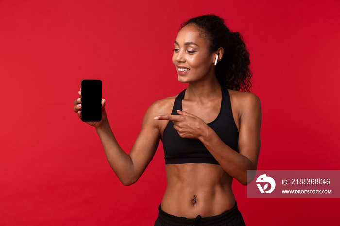 Portrait of healthy african american woman in black sportswear using smartphone and earbuds, isolate