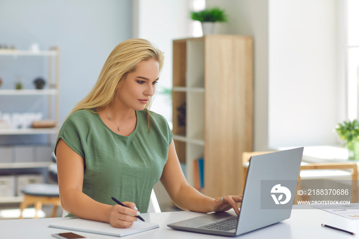 Serious young good-looking blonde woman sitting at table with laptop computer and using online learn