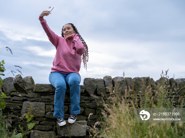 Teenage (16-17) girl sitting on stone wall�taking selfies