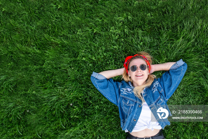 戴着墨镜、穿着牛仔夹克的快乐女孩躺在绿草上，看着镜头。