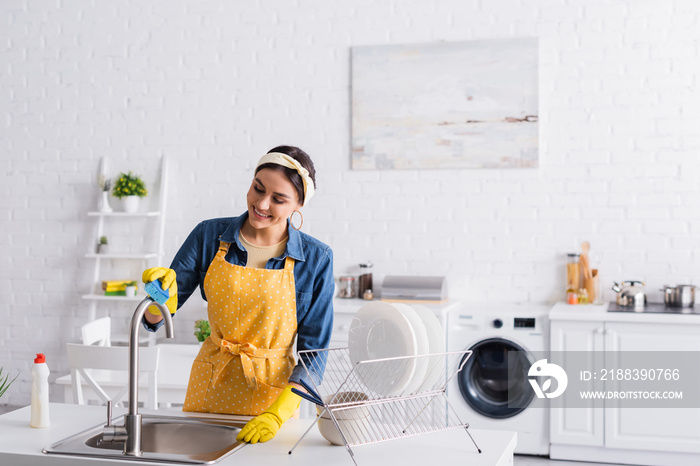 Smiling housewife cleaning faucet near plates in kitchen