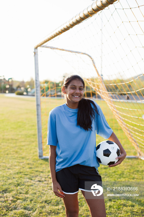 Portrait of smiling soccer player holding ball while standing on grassy field against clear sky