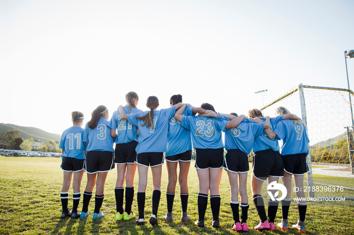 Rear view of girls in soccer uniforms standing on grassy field against sky