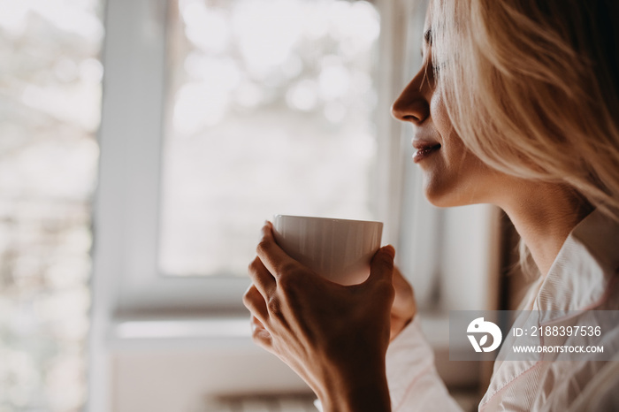 Woman holding a cup of coffee, sitting by the window in the morning.