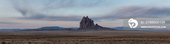 Dramatic panoramic landscape view of a dry desert with a mountain peak in the background during a vibrant cloudy sunrise.Taken at Shiprock, New Mexico, United States.