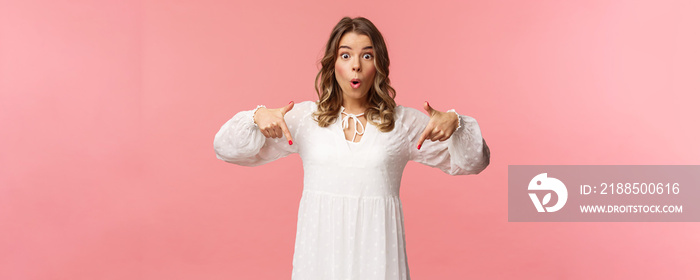 Waist-up portrait of interested, excited attractive blond 20s woman showing person something really cool, say wow pop eyes camera excited, pointing fingers down at great offer, pink background