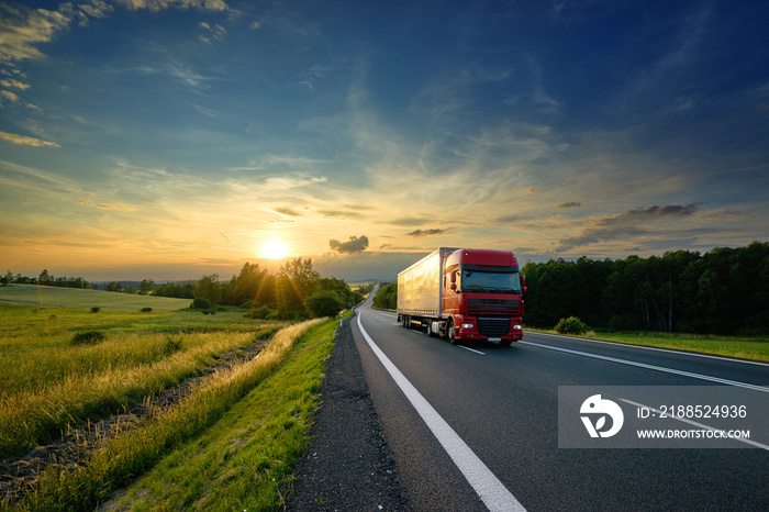 Red truck driving on the asphalt road in rural landscape at sunset