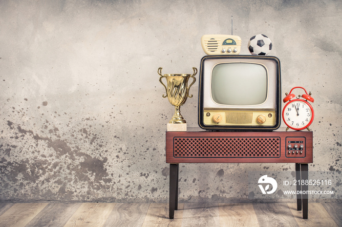 Retro outdated television from 50s, soccer ball, golden winner cup, radio receiver, orange alarm clock on wooden TV stand front aged textured concrete wall background. Vintage old style filtered photo