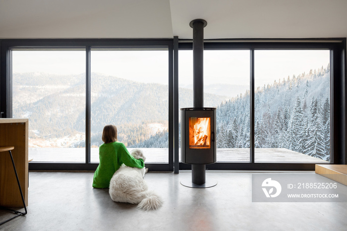 Woman sitting with dog near fireplace and panoramic window at modern living room with stunning view on snowy mountains. Concept of rest in houses or cabins on nature. Idea of escape from everyday life