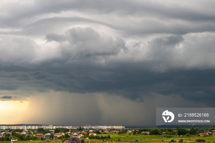 Moody landscape with dark stormy clouds with falling heavy downpour shower rain over distant town buildings in summer.