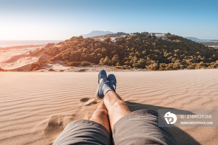 feet of a traveler in shorts and hiking shoes on the background of a paradise landscape with golden sand and blue sea in the background. Wellbeing and exploration concept